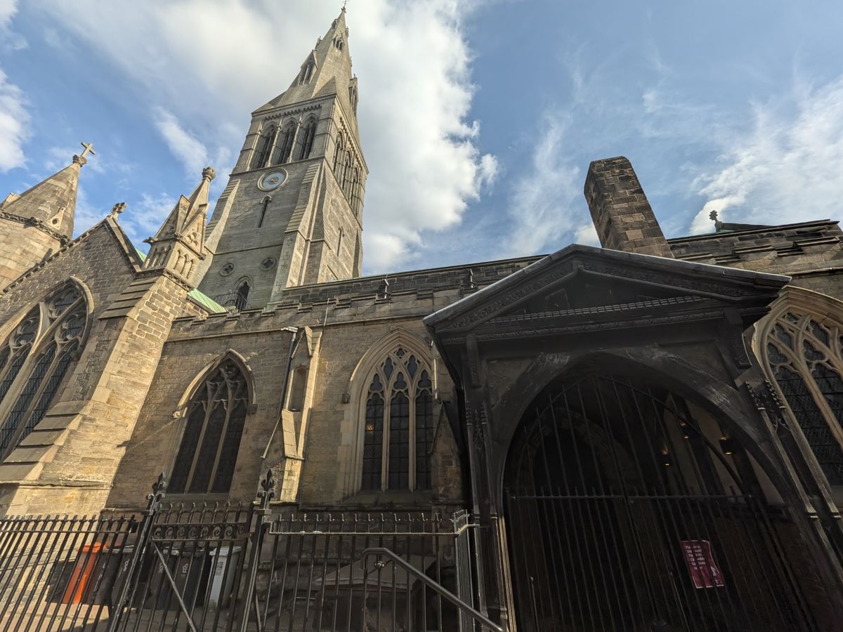 Low-angle view of a historic stone church and tall bell tower against a bright blue sky with scattered clouds.