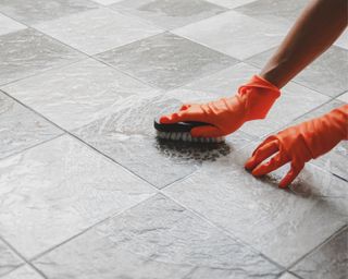 Tiled bathroom floor being cleaned by someone in orange gloves, using scrubbing brush and soapy solution