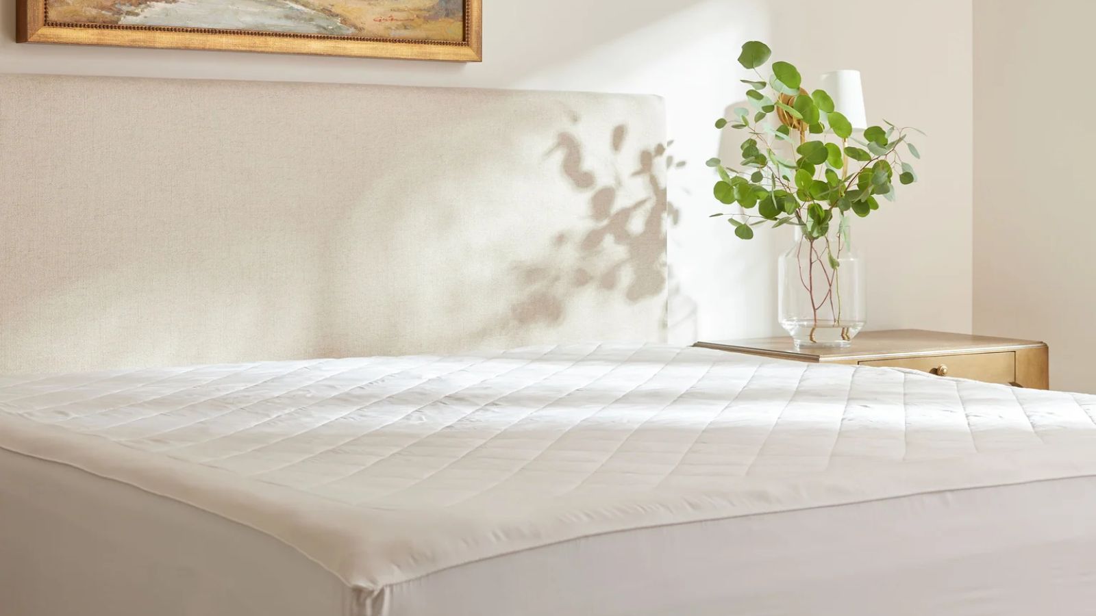 Cozy Earth bamboo mattress pad shown on a white bed in a white bedroom, with an artwork in gold frame above, and a wooden nightstand with a glass vase with green plant and white lamp.