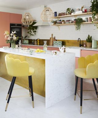 A galley kitchen with kitchen island with mustard yellow velvet bar stools, pink cabinets and open shelving