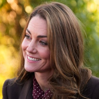 A headshot of Kate Middleton wearing a maroon coat and dress and smiling in front of yellow and green trees