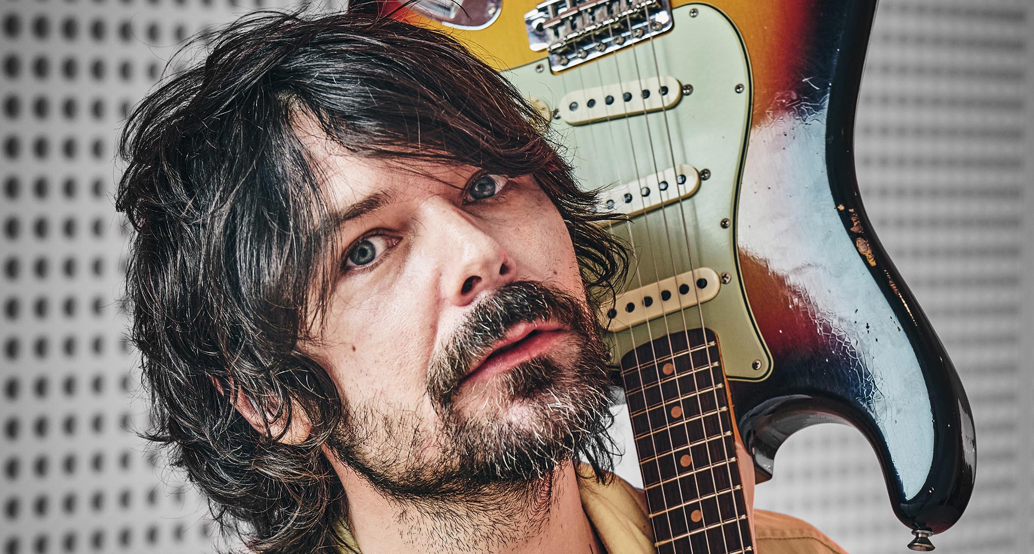 Simon Neil of Biffy Clyro wears a custard-colored jacket and green T-shirt and poses with a sunburst Fender Stratocaster