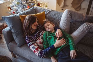 A woman hugging a sleeping young boy on a sofa in front of a Christmas tree