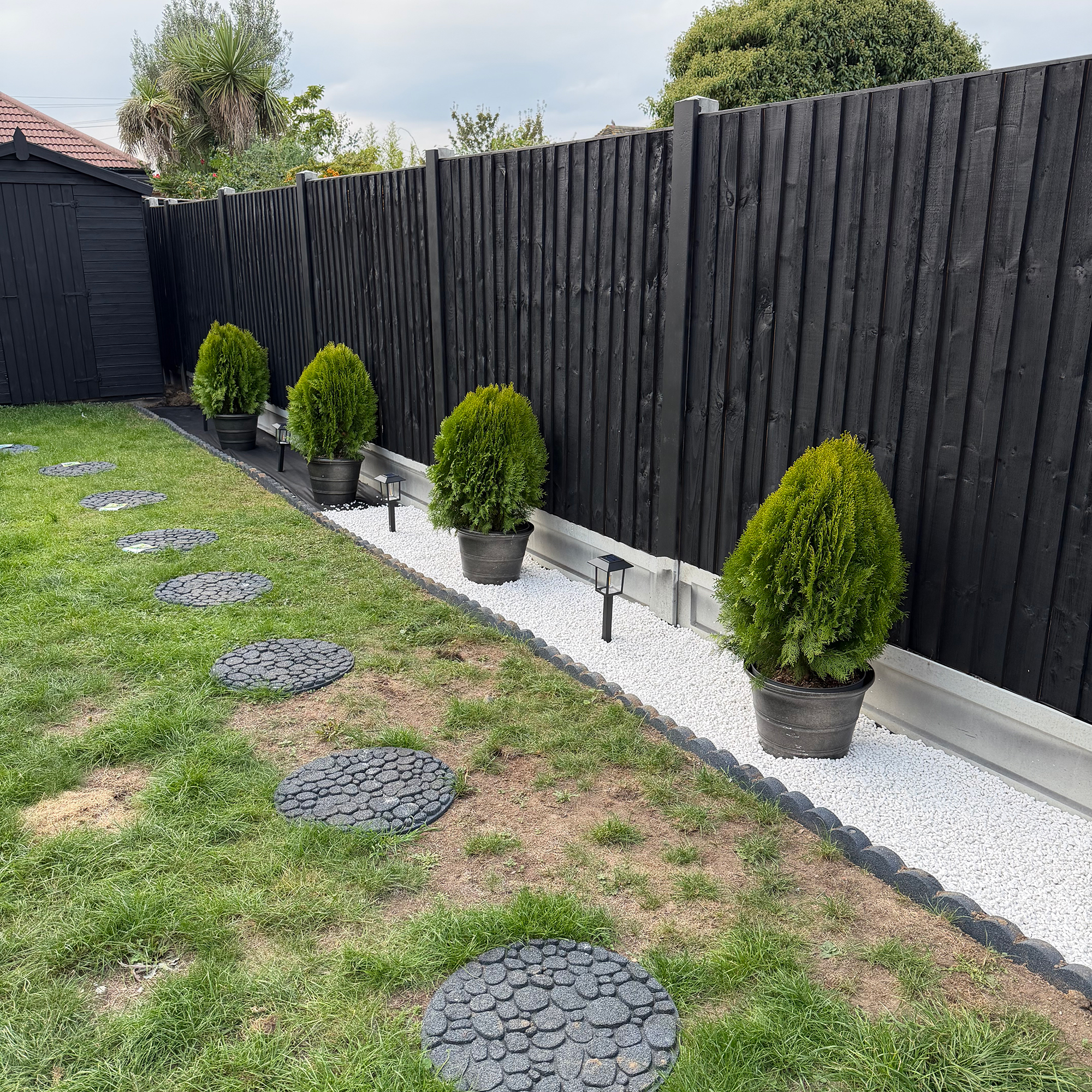 Garden with round pavers in grass, potted small trees by black fence and small lights in ground