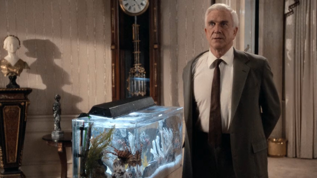 Leslie Nielsen looking guilty with his hands behind his back while standing next to a fish tank.