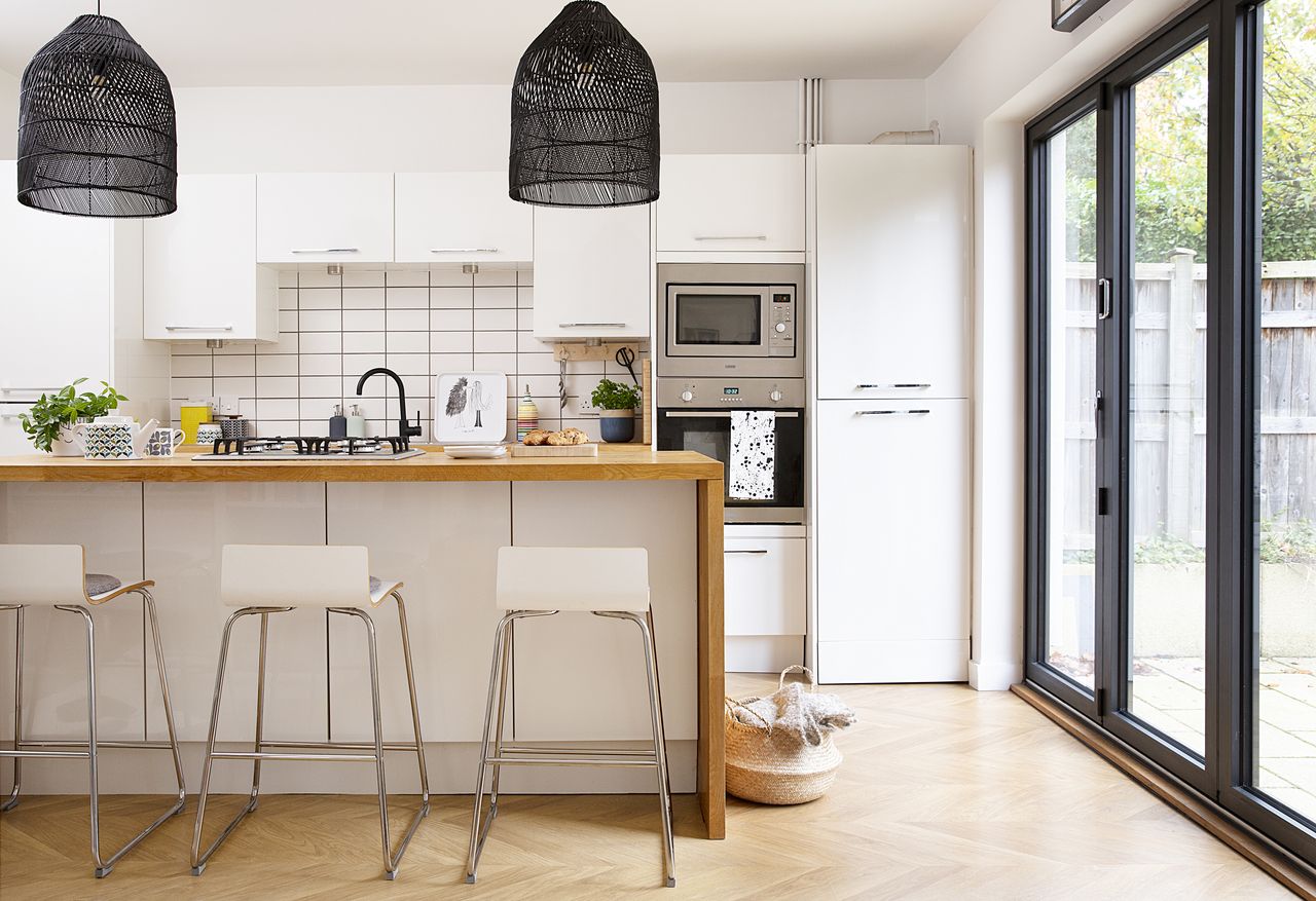 Kitchen in the open-plan kitchen-diner with white units and wall hung cabinets, an island with wrap-around wooden worktop, white bar stools and black mesh pendant lights