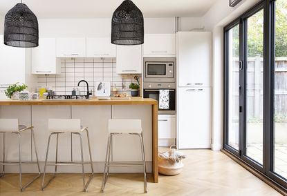 Kitchen in the open-plan kitchen-diner with white units and wall hung cabinets, an island with wrap-around wooden worktop, white bar stools and black mesh pendant lights