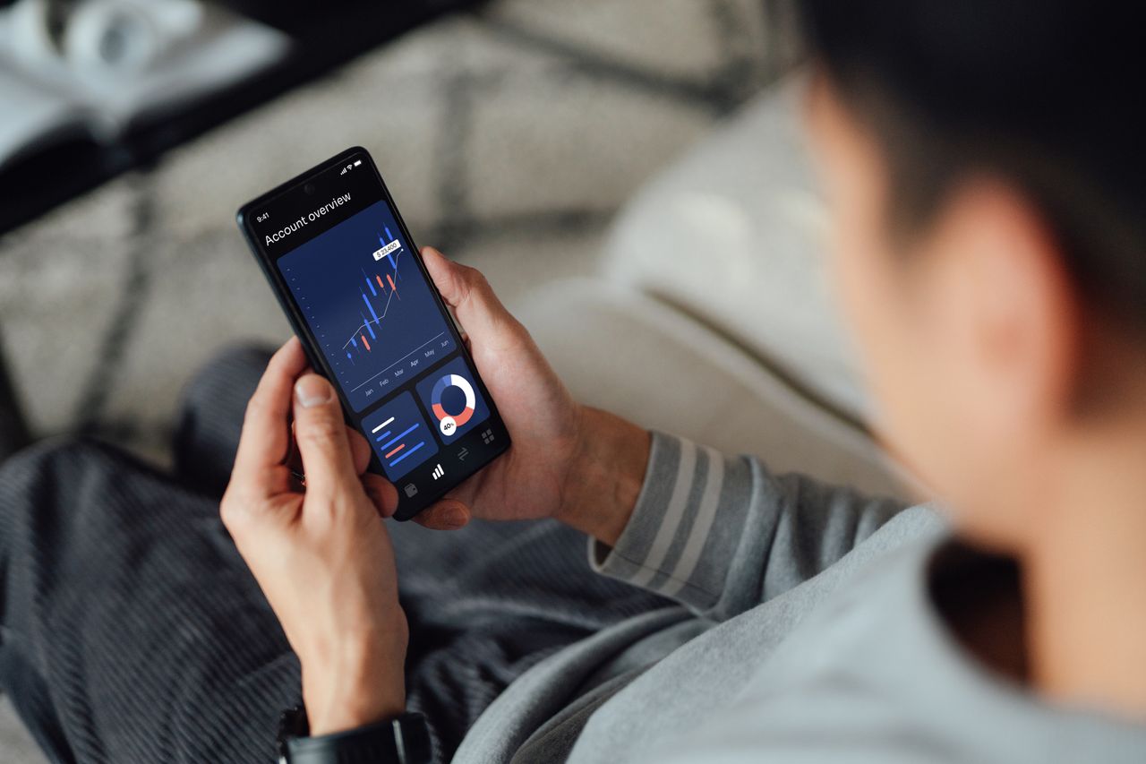 Over the shoulder view of young man checking financial data with mobile banking app on smart phone while sitting on sofa at home