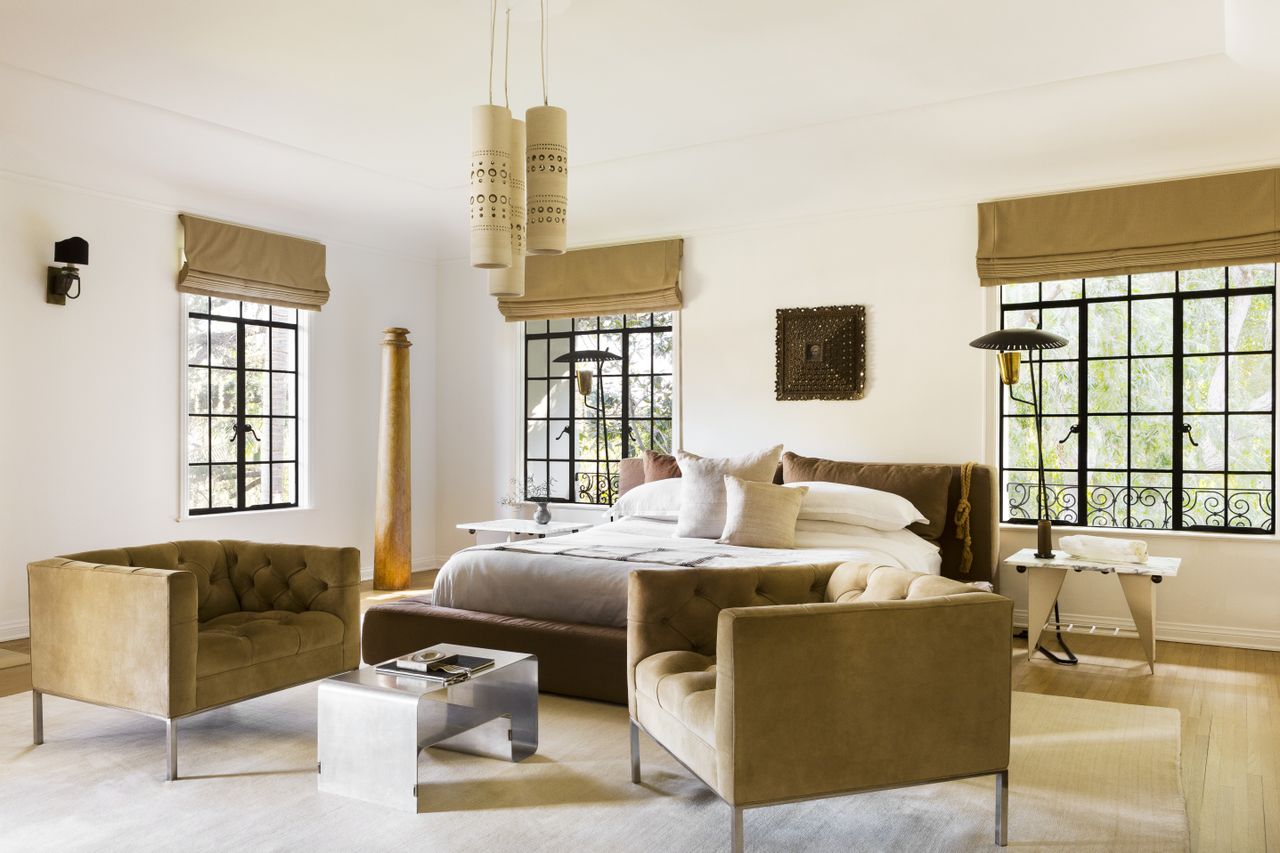 A modern bedroom with white walls and ceiling and wood flooring, with two dark beige velvet armchairs facing each other at the foot of the bed with a headboard of the same material, matching the roman brands hung above the black-framed windows