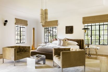 A modern bedroom with white walls and ceiling and wood flooring, with two dark beige velvet armchairs facing each other at the foot of the bed with a headboard of the same material, matching the roman brands hung above the black-framed windows