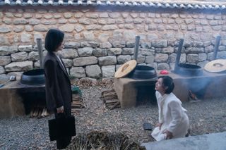 a woman (Song Hye-kyo as Moon Dong-eun) stands while another woman (Cha Joo-young as Choi Hye-jeong) begs on her knees, in front of a set of traditional Korean cauldrons, in Netflix k-drama 'The Glory'
