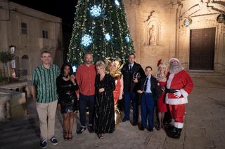 The cast of The Madame Blanc Mysteries stand around the brightly lit and decorated Christmas tree in the town square: Niall (Aonghus Weber), Celine (Margeaux Lampley), Dom (Steve Edge), Jean (Sally Lindsay), Gloria (Sue Vincent), who is dressed as a Christmas cracker, Caron (Alex Gaumond), Gendarme Richard (Jacqueline Berces), Judith (Sue Holderness) and Jeremy (Robin Askwith) who is dressed as Santa