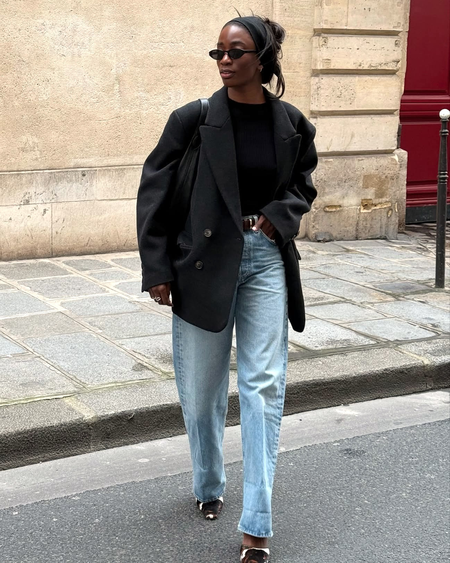 Style influencer Liv Madeline crosses a street in Paris, France wearing black oval sunglasses, a black headband, an oversize black blazer, a black top, belt, light-wash barrel jeans, and cow-print mule heels.