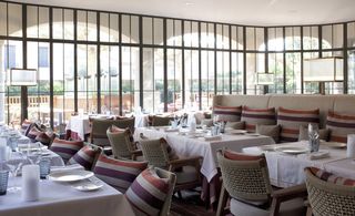 Rivea, Saint Tropez, France. A restaurant with rows of white decorated tables, chairs with striped cushions, square floor lamps and floor to ceiling windows.