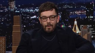 Jorma Taccone wearing all black with glasses guesting on Late Night with Seth Meyers