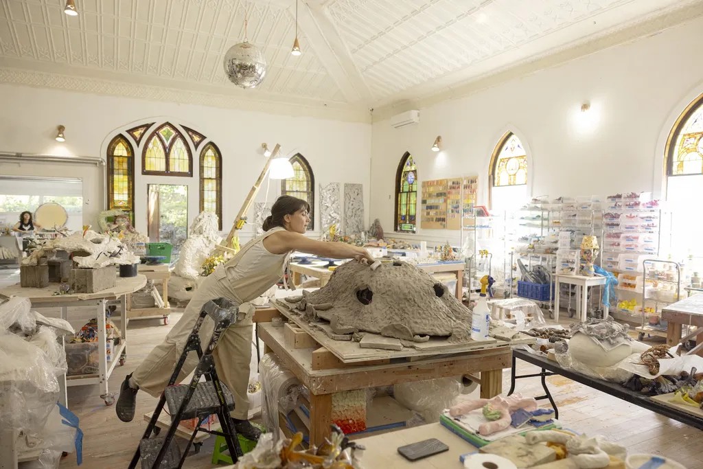 A young woman dressed in pale beige dungarees in an objects-filled artist studio works on a ceramic sculpture of a big creature in a naturally lit space.