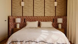 Image of a red-and-white patterned extended headboard in a beige room where one wall is upholstered. The bed has white bedding with black stitching around the bottom.