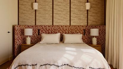 Image of a red-and-white patterned extended headboard in a beige room where one wall is upholstered. The bed has white bedding with black stitching around the bottom.