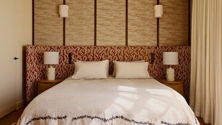 Image of a red-and-white patterned extended headboard in a beige room where one wall is upholstered. The bed has white bedding with black stitching around the bottom.