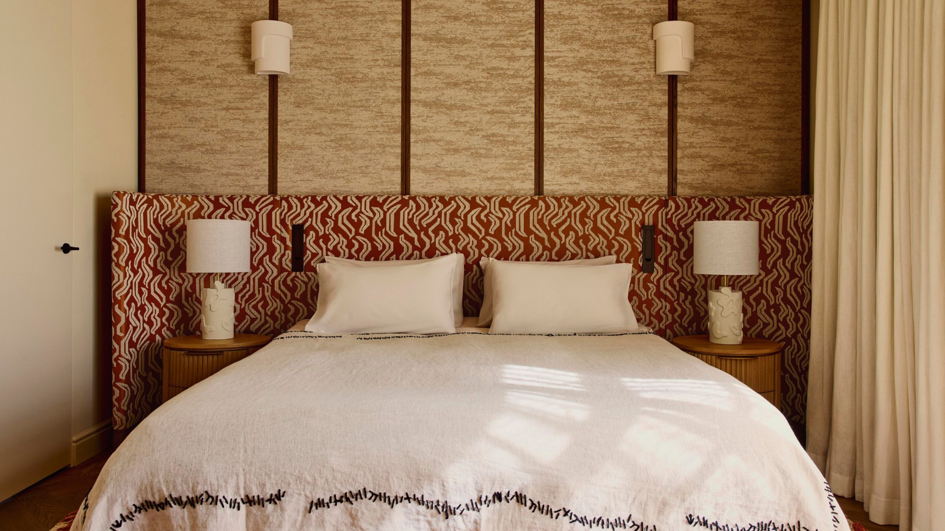 Image of a red-and-white patterned extended headboard in a beige room where one wall is upholstered. The bed has white bedding with black stitching around the bottom.