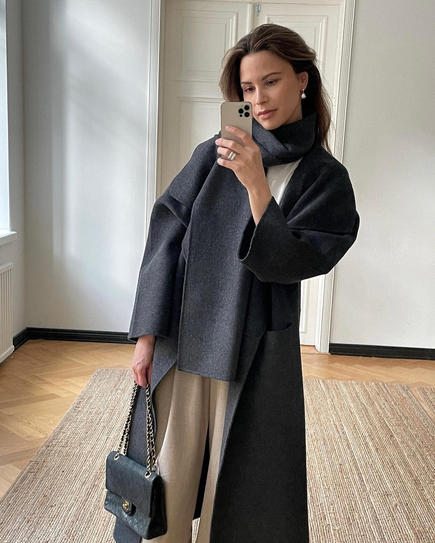 Finnish style influencer poses for a a mirror selfie wearing a gray scarf coat, white tee, and tan pants