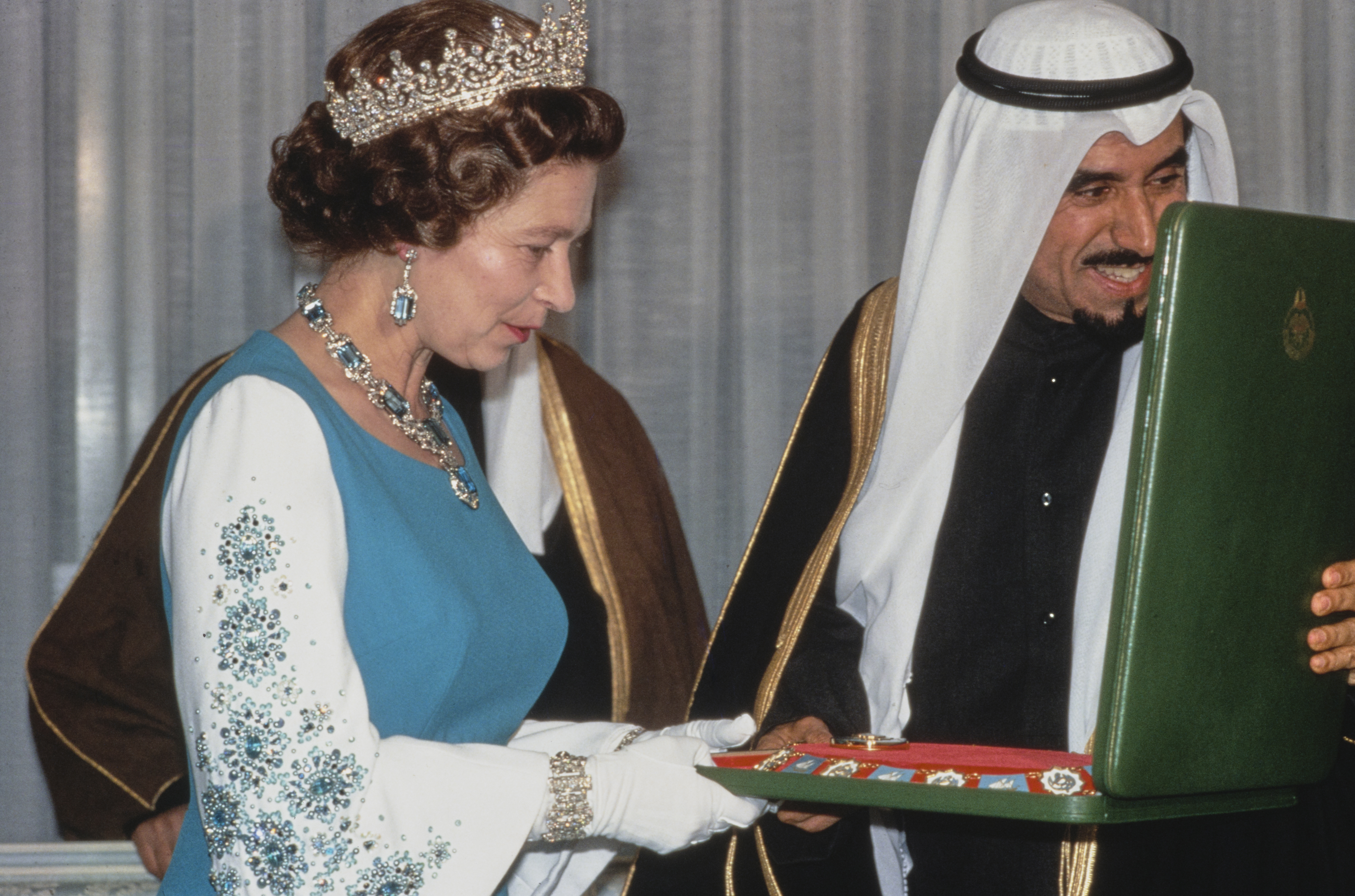 Queen Elizabeth wearing a tiara opening a box with a necklace inside given by a man in an Arab head dress