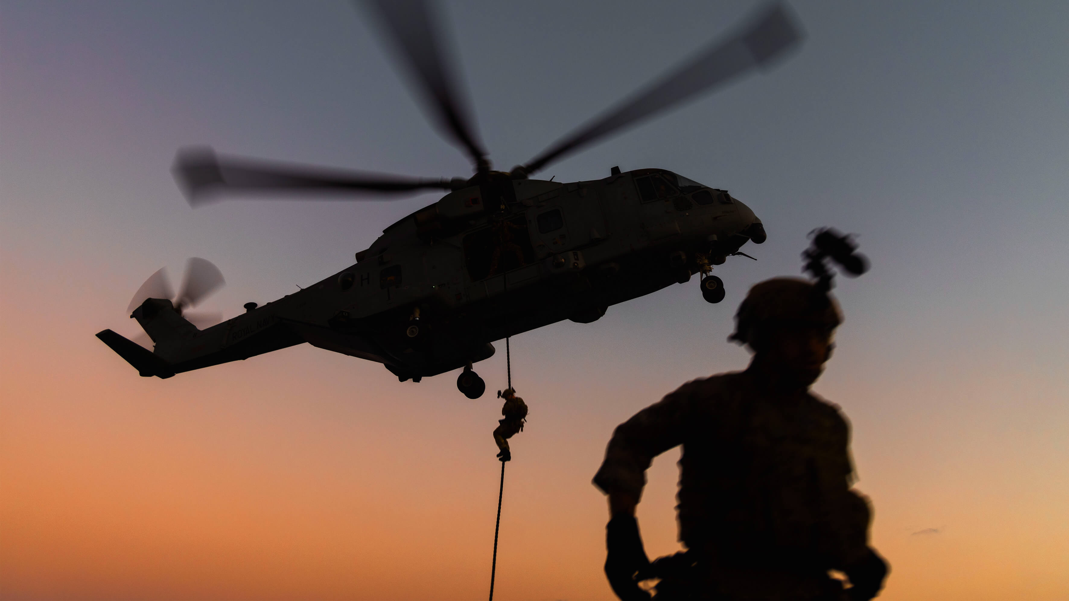 Soldier silhouetted against helicopter flying above. Another soldier rappels from helicopter. 