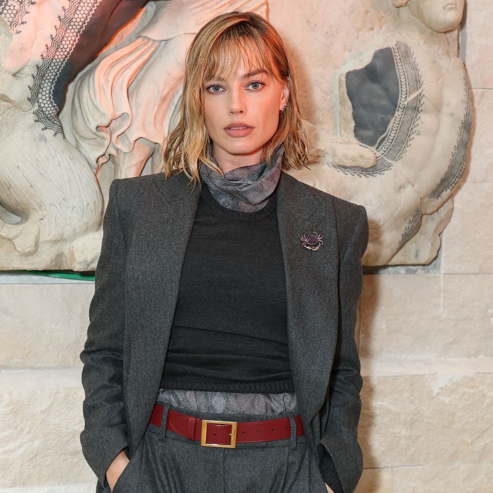Margot Robbie wears burgundy flats with grey wool trousers and a grey longline blazer.