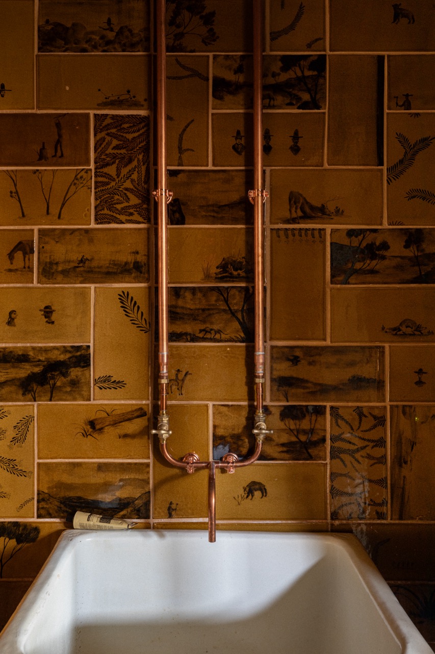 A white sink with copper fixtures against amber tiles with hand-painted drawings across a backsplash.