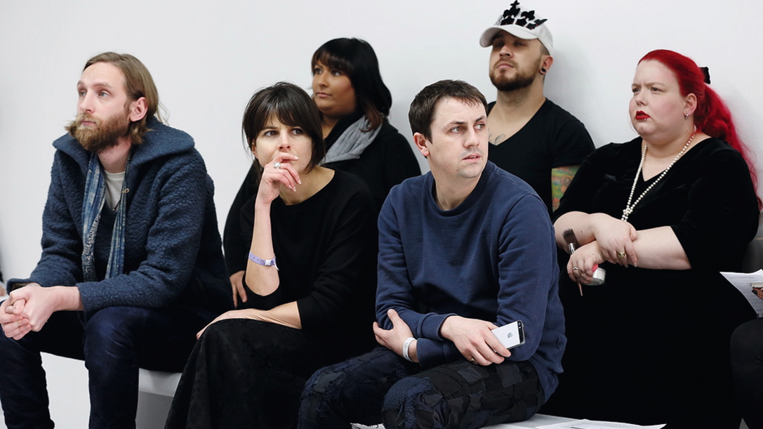Six people, all wearing black, sit on benches and concentrate at a casting at London Fashion Week