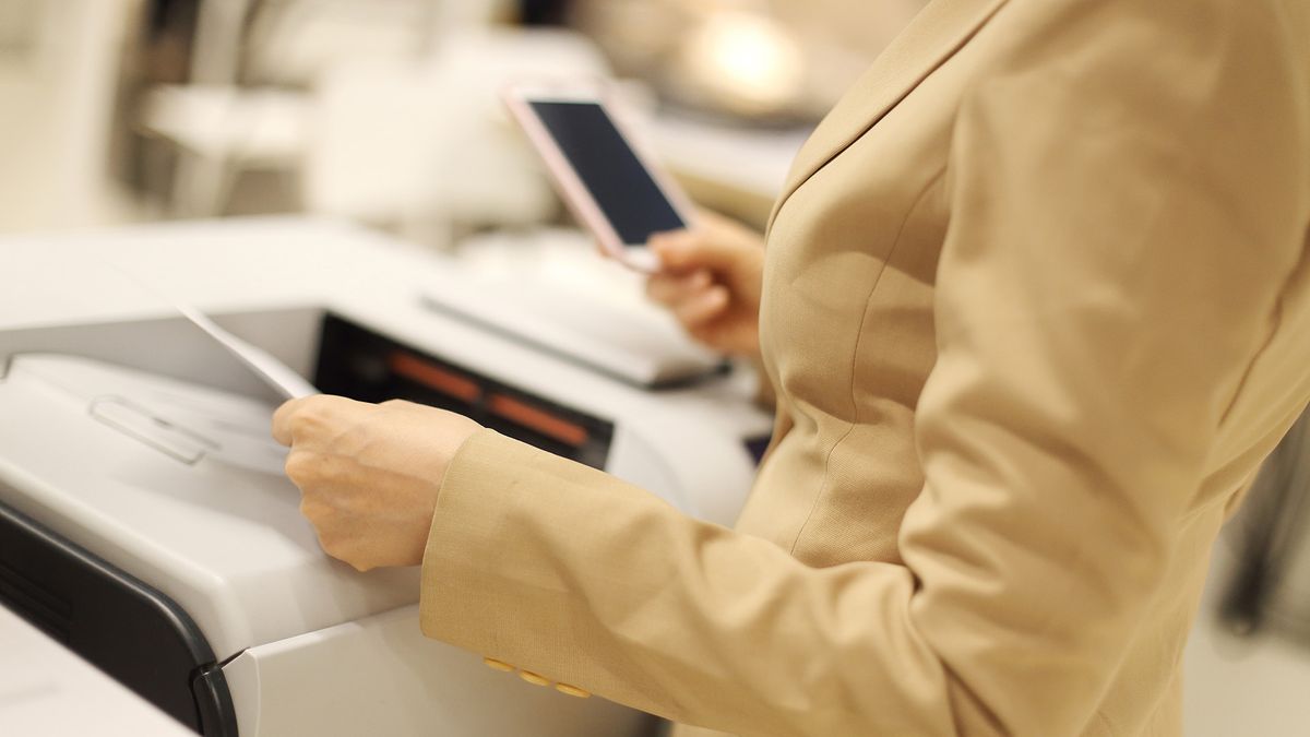 Woman standing at office printer holding an iPhone in one hand.