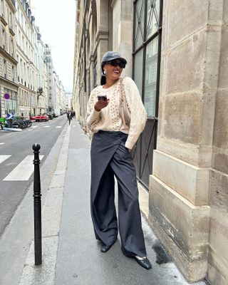 French style influencer Lena Farl smiles and poses on the streets of Paris wearing a faded baseball cap, statement earrings, trousers, sock, and ballet flats