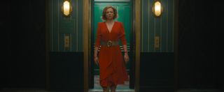 Sheila (Sarah Snook) exits a hotel elevator and walks down a hotel corridor, wearing a red dress with a thick brown leather belt around her waist, and looking concerned