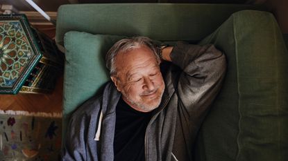 Smiling senior man with hand behind head napping on sofa at home