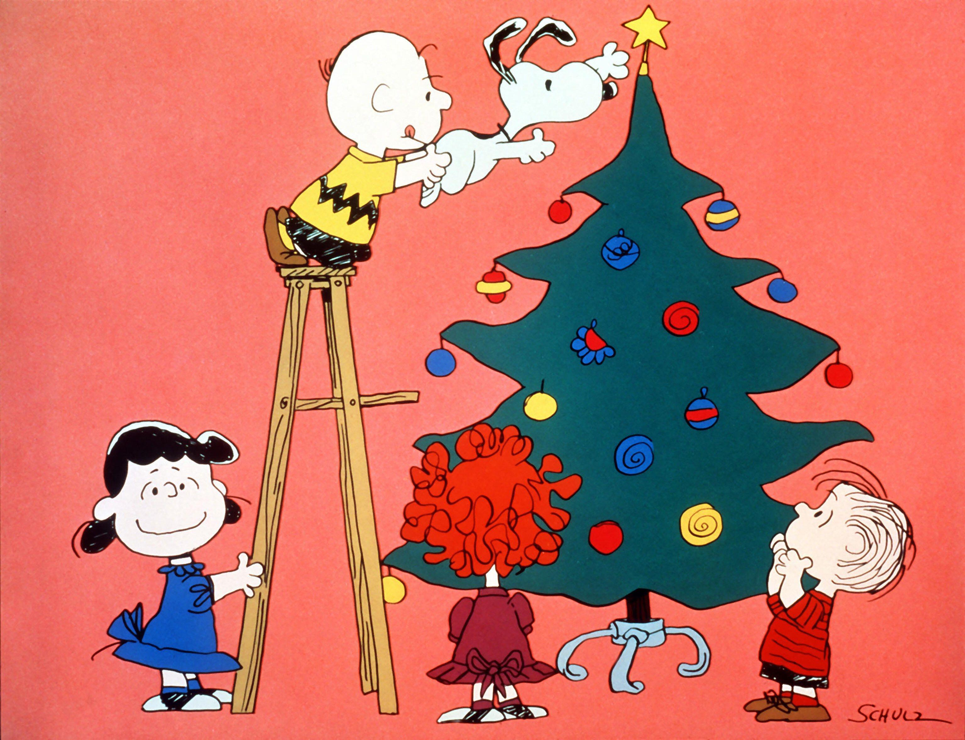 Charlie Brown stands on a wooden ladder holding Snoopy up to place a star on a Christmas tree, while Lucy, Sally and Linus watch against a pink background.