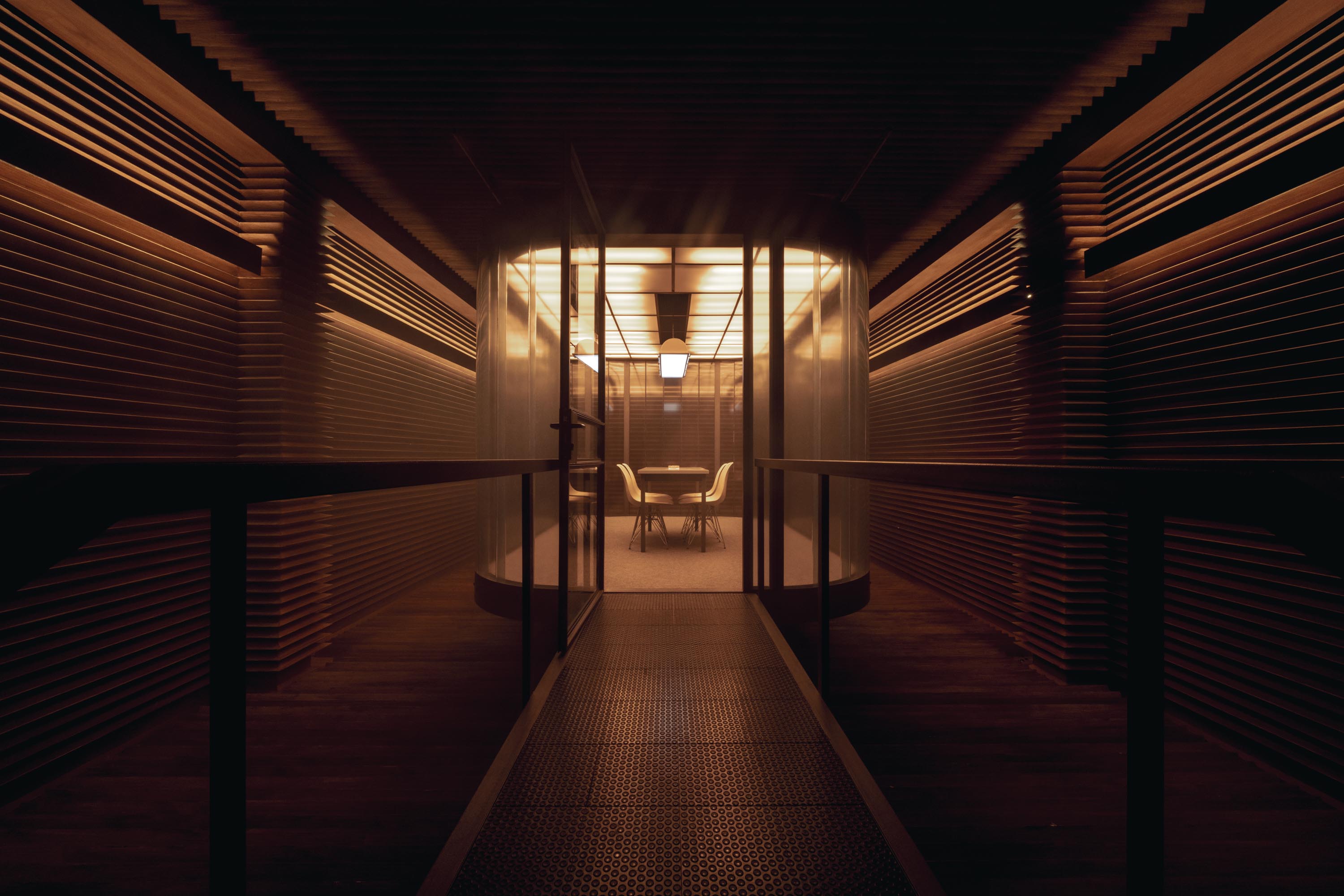 A digital rendering of a secret booth crafted from wood, Plexiglass, and steel shows a warmly lit cubicle room with a central desk and set of chairs, illuminated by back-lit ceiling panels, while two handmade sketches of rooms capture their interiors from above.