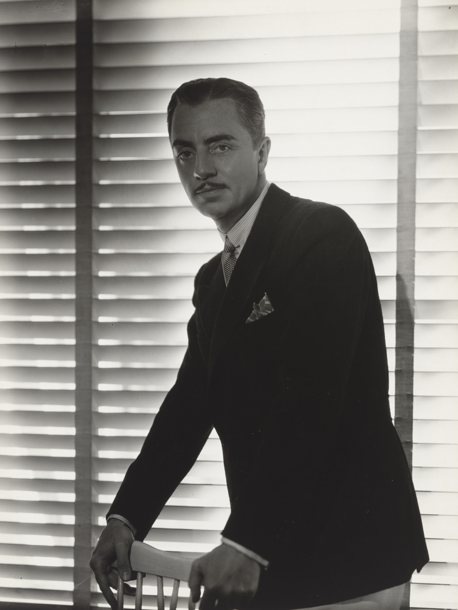 William Powell, with a thin moustache, wears a dark jacket with a patterned pocket square and leans on the back of a wooden chair in front of a brightly backlit window blind.