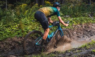 A white, male cyclist riding a gravel bike through a deep, muddy puddle, with water splashing out from the side