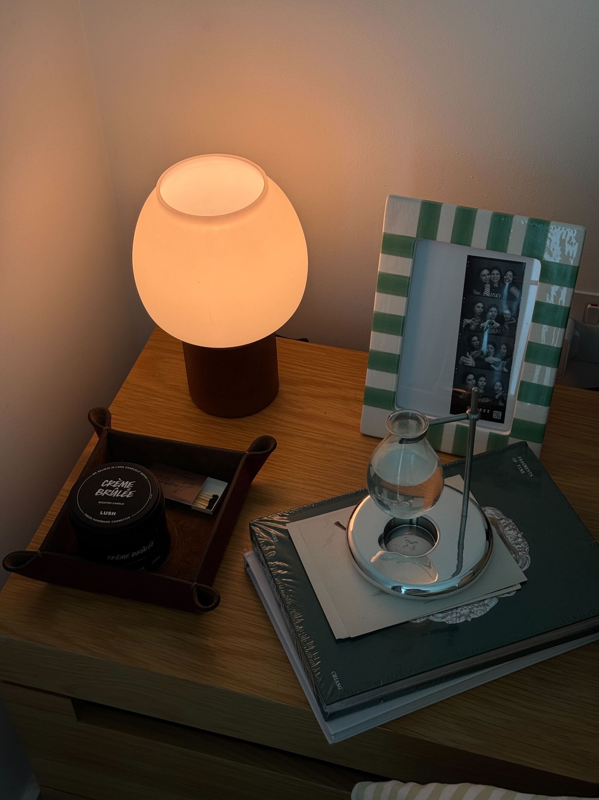 A wooden nightstand with a small red and white table lamp, a leather catch-all, a stack of books, a diffuser, and a green striped picture frame.