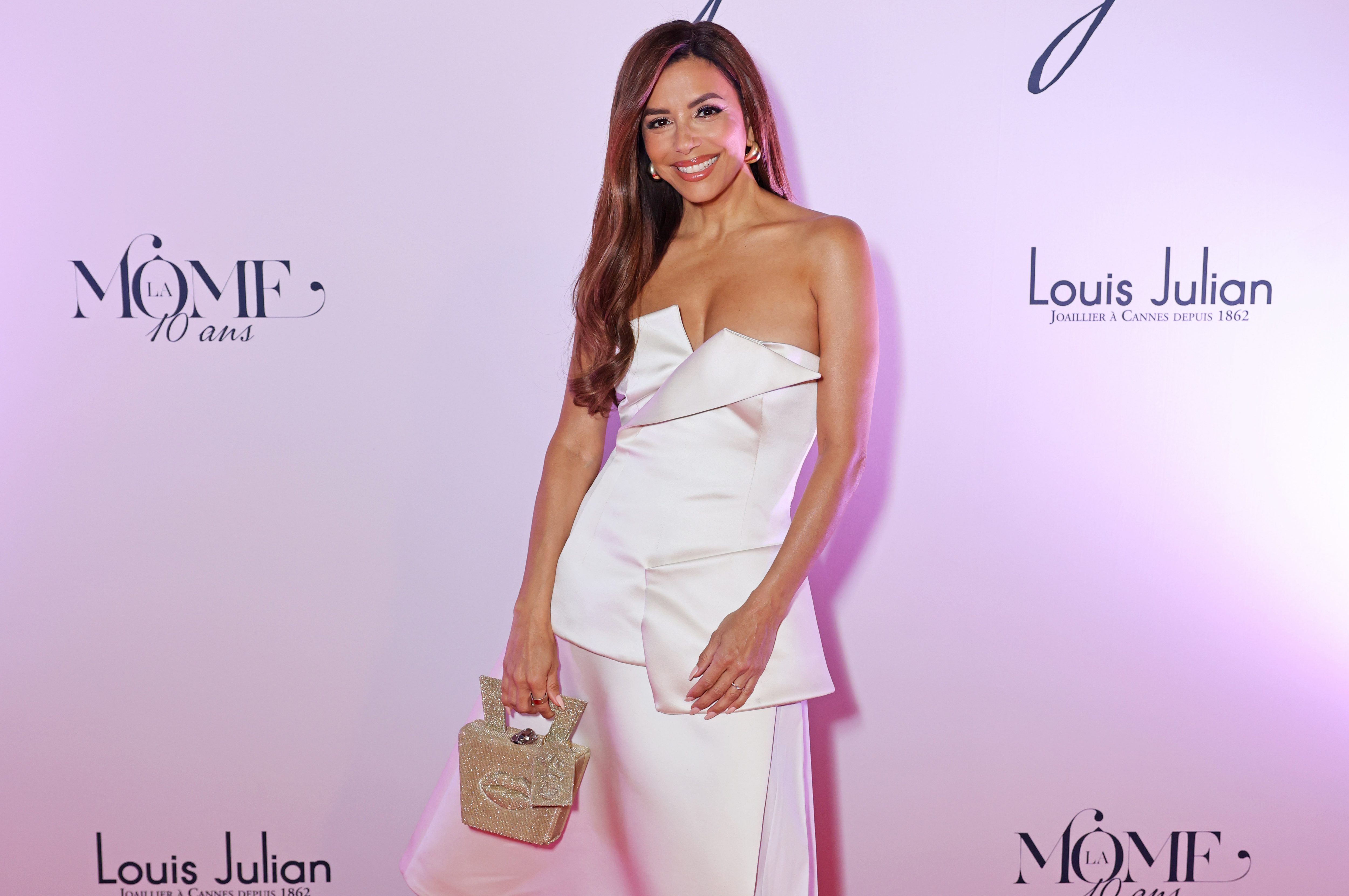Wide shot with Eva Longoria wearing white asymmetrical dress, gold jewelled handbag, and glossy, curled hair. Eva Longoria attends La Môme 10 year anniversary dinner celebration on May 17, 2025 in Cannes, France.