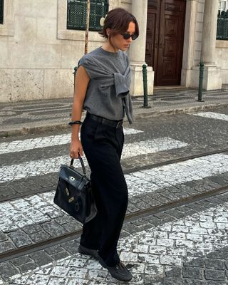 fashion influencer Debora Rosa crossing the street wearing a hair claw clip, black oval sunglasses, a sleeveless gray sweater with a built-in scarf, black belt, black trousers, Hermes bag, and black moccasin loafers