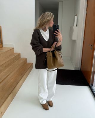 Anouk Yve wearing a white T-shirt, brown sweater, white jeans, a tan bag, and tan loafers.