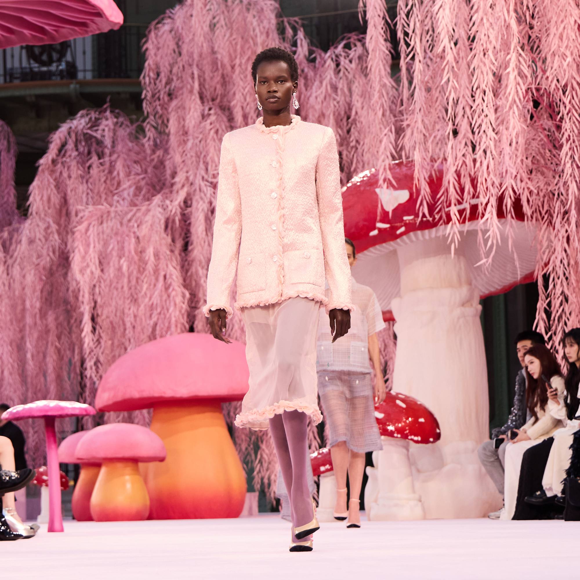 A model walks the runway surrounded by oversize mushrooms and pink leaves