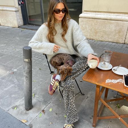 Fashion influencer @lunaisabellaa sitting outside at a cafe in Barcelona with her dog in her lap, wearing a chic spring outfit with a soft grey sweater, leopard print jeans, and Adidas SL 72 sneakers.