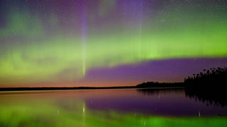 The Northern Lights dance over a lake with a faint orange from the sunset on the horizon