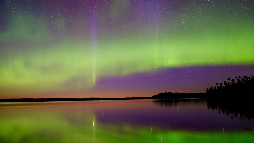 The Northern Lights dance over a lake with a faint orange from the sunset on the horizon