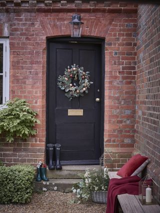 Christmas exterior with black painted door, wreath, wellies on steps, bench with red velvet cushion and throw, lanterns, basket with fresh foliage