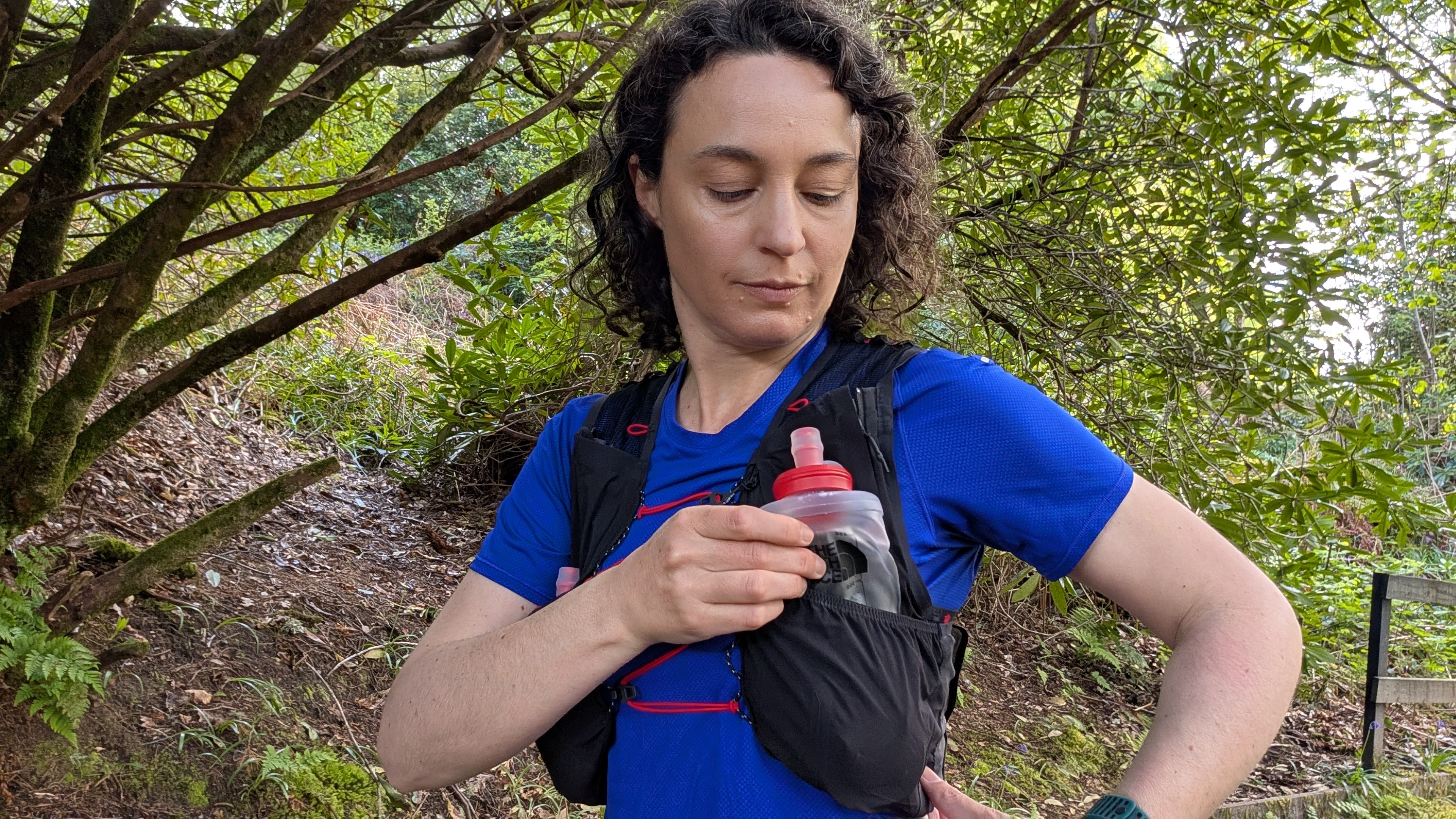 A woman trail runner in the woods wearing a blue t-shirt pulls a water bottle from a black hydration vest from The North Face