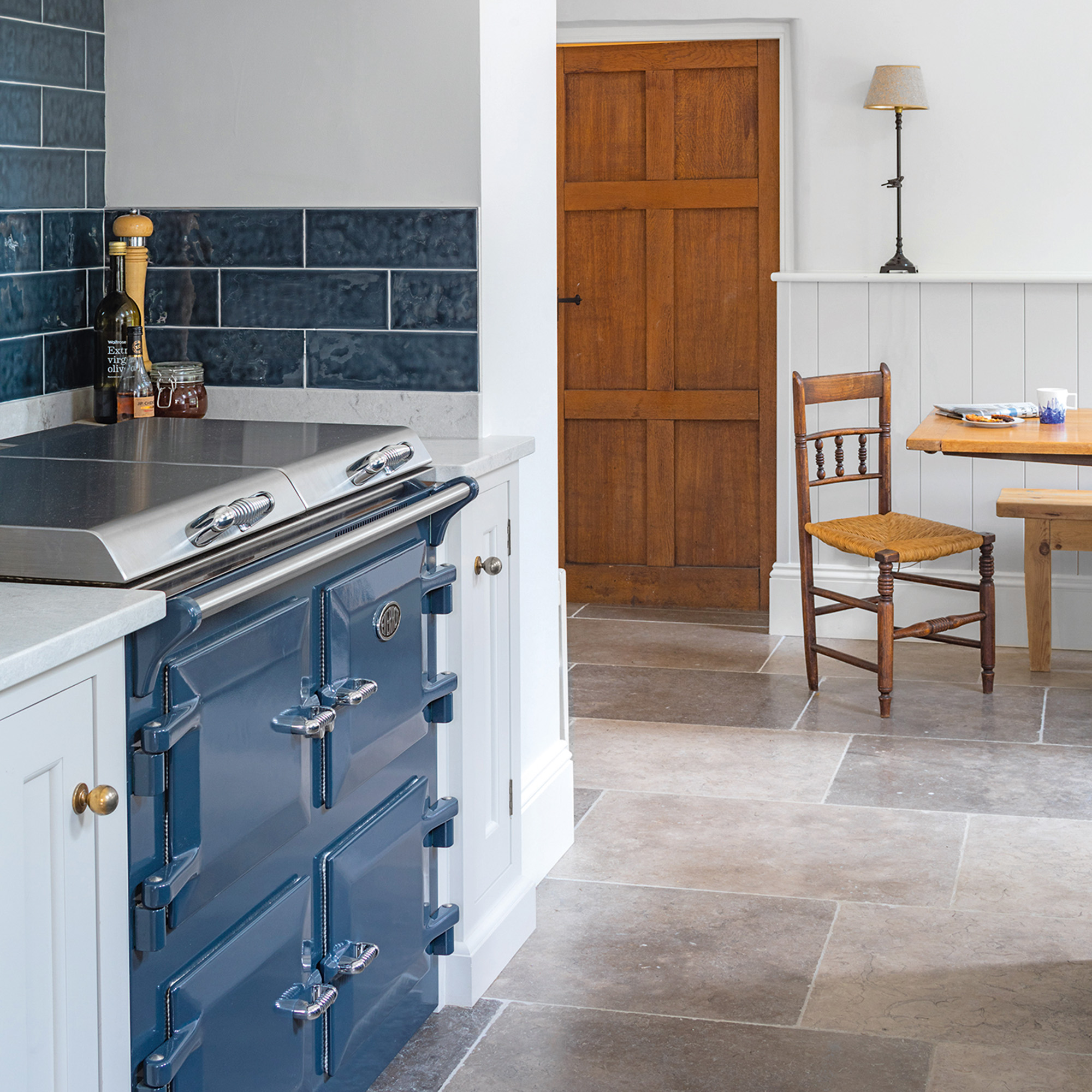 kitchen with white walls and blue range cooker with navy metro tile splashback and grey stone flooring unpainted wooden door on back wall beside wooden table and chairs