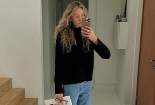 Woman wearing black cashmere sweater and jeans, taking mirror selfie.